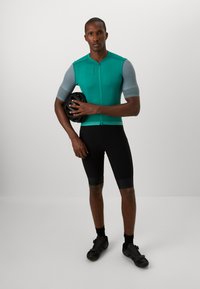 Maglietta da ciclismo a maniche corte color teal con maniche grigie testurizzate, abbinata a pantaloni corti da ciclismo neri e scarpe, mentre tiene un casco nero.