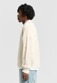 Cream-colored fluffy sweater with dropped shoulders and ribbed cuffs. Features a small metallic detail on the sleeve, worn over a white shirt.