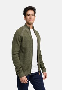 Cardigan zippé vert en tissu tricot texturé. Doté d'un col montant et de poignets côtelés, il est associé à un T-shirt blanc et à un jean foncé.