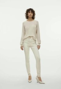 Beige garais piedurkņu tops, kas savienots ar gaiši beidzot skinny džinsiem un baltām augstpapēžu kurpēm. Apģērbs izceļas ar vienkāršu, minimālistisku dizainu un smalkām tekstūrām.