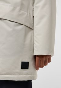 Veste imperméable gris clair avec une texture lisse, présentant un logo en forme de patch noir au bas et un design décontracté.