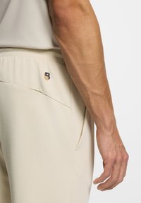 Beige joggers featuring a textured fabric, elastic waistband, and side pockets. Small black and gold logo on the rear pocket.