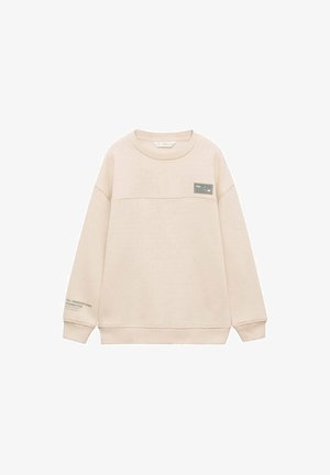 Sweatshirt beige clair avec un col rond, des poignets et un ourlet côtelés. Présente une étiquette verte sur la poitrine et un détail textuel sur la manche.