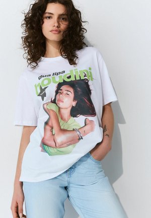T-shirt blanc avec un imprimé graphique coloré d'une personne en haut vert. Le t-shirt a une coupe décontractée et des manches courtes. Jeans en denim clair.