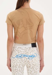 Beskåret beige T-shirt med kranietryk og røde detaljer, parret med hvide leopardmønstret jeans med blåt logo på baglommerne.