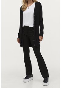 Kvinna i vit blouse, lång svart knäppt cardigan, svarta byxor och svarta plattformssneakers som står mot en neutral bakgrund.