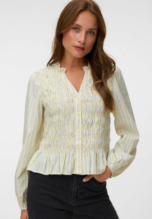Femme aux longs cheveux bruns portant un chemisier rayé jaune et blanc avec des boutons et des détails smockés, debout avec une main dans la poche d’un pantalon noir.