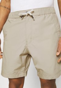 Man wearing beige drawstring shorts and white top, with a visible Roman numeral tattoo on upper left arm.