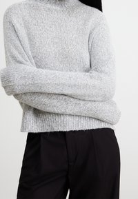 Grijze gebreide trui met een hoge kraag, een losse pasvorm en een geribbelde textuur. Gecombineerd met zwarte broeken, wat zorgt voor een casual gelaagde stijl.