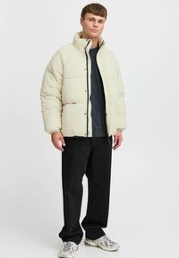 Puffjakke i lys beige med høj krave, lynlås og quiltet design. Parret med mørke bukser og sneakers.