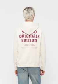 Cream-colored hoodie with a front kangaroo pocket, featuring bold burgundy text and graphics on the back. Made of soft fabric with a textured finish.