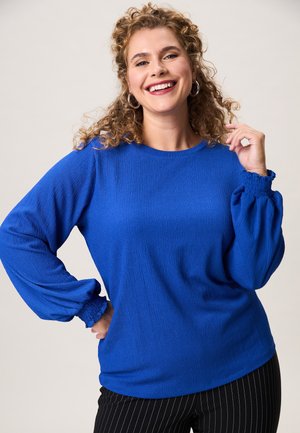 Lachende vrouw met krullend haar draagt een blauw shirt met lange mouwen en zwarte broek met krijtstreepjes, poseert met hand op heup en opgeheven vingers.