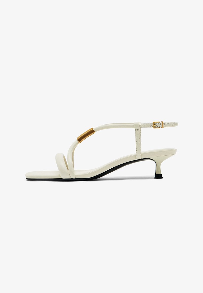 Sandale blanche à talon bas avec des fines brides, une boucle dorée et un accent doré sur la bride avant, vue de profil.