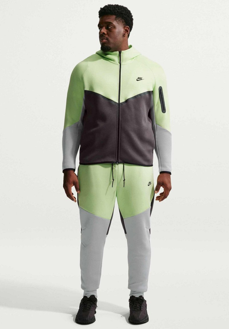Mand står iført neongrøn, grå og sort Nike træningsdragt med hættetrøje og matchende joggers, kigger mod venstre.