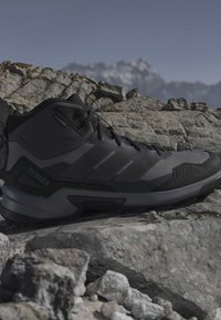 Μαύρο και γκρι παπούτσι πεζοπορίας Adidas Terrex σε βραχώδες έδαφος με θολό ορεινό όγκο στο φόντο κάτω από καθαρό ουρανό.
