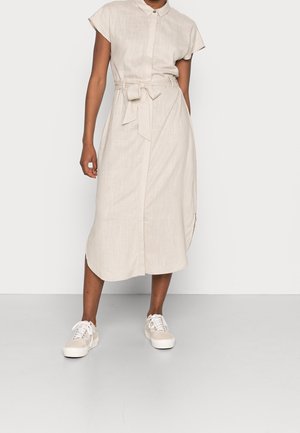 Personne portant une robe midi beige à manches courtes avec ceinture à la taille et col boutonné, associée à des baskets beiges et blanches.