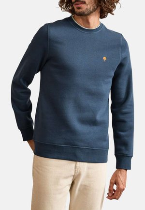 Homme portant un sweat-shirt à col rond bleu avec un petit logo d'arbre orange sur la poitrine et un pantalon beige, debout avec les mains détendues.