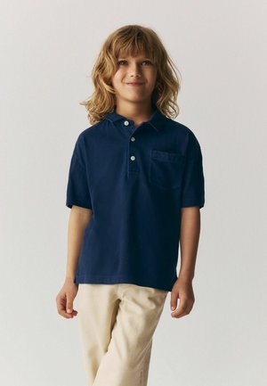 Kind mit schulterlangem blonden Haar, das ein dunkelblaues Poloshirt und beige Hosen trägt, vor einem schlichten hellen Hintergrund stehend.