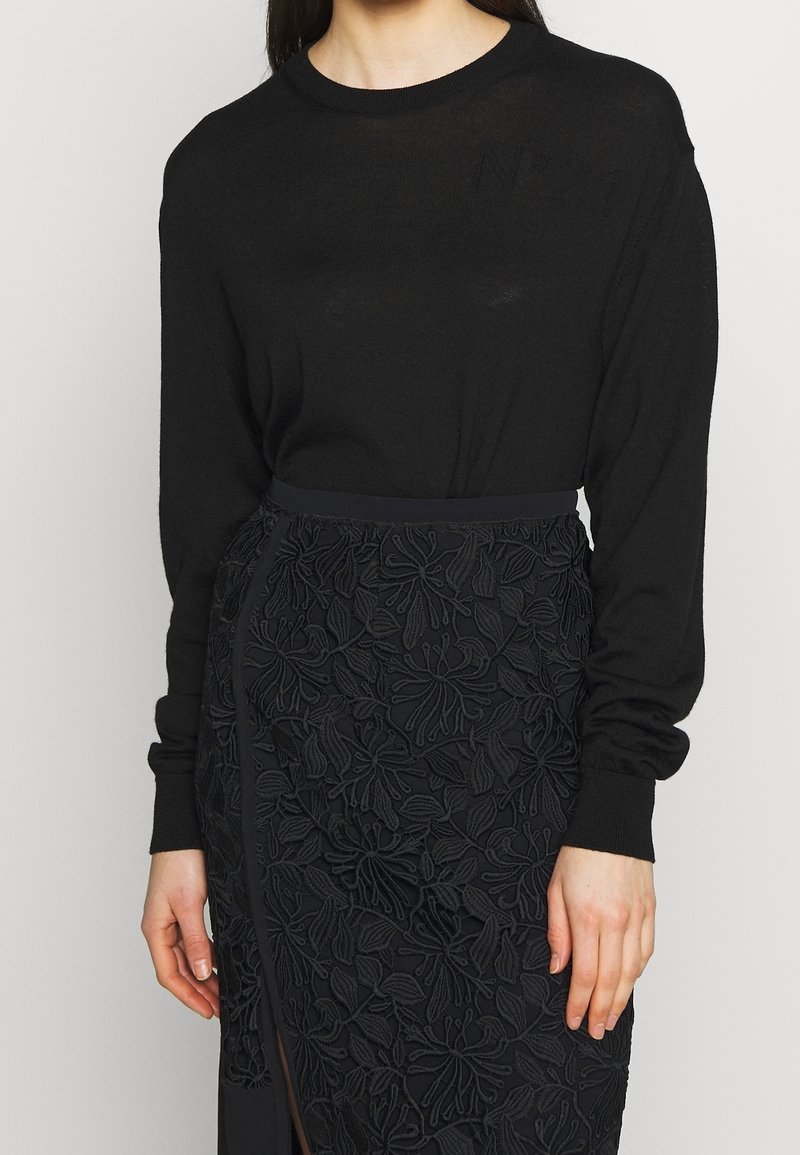Femme portant un pull noir à manches longues et une jupe noire avec une broderie florale et une fente à l’avant.