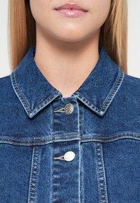 Τζιν μπουφάν με μπλε ύφασμα, που διαθέτει δομημένο γιακά, ασημένια κουμπιά και ορατές λεπτομέρειες ραφής. Η υφή φαίνεται ομαλή.