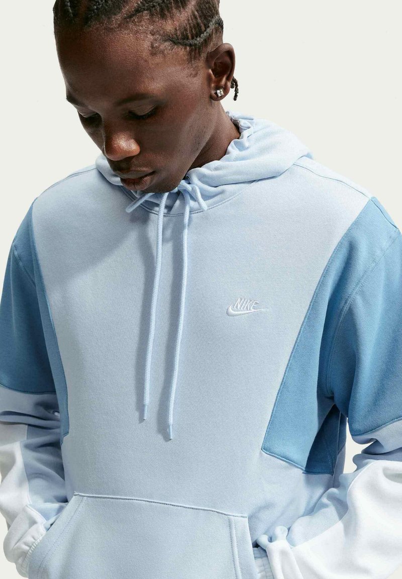Persoon met gevlochten haar kijkt naar beneden, draagt een lichtblauwe Nike-hoodie met handen in de zakken tegen een egale lichte achtergrond.