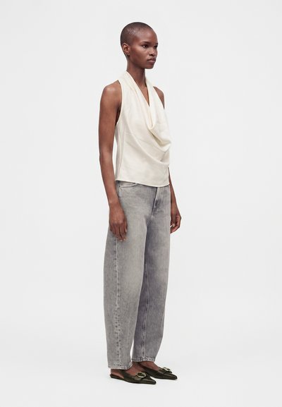 Femme aux cheveux courts portant un haut sans manches blanc drapé, un jean gris à jambes larges et des ballerines noires pointues avec boucles.