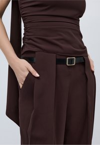 Γυναίκα ντυμένη με σκούρο καφέ φόρεμα με πιέτες στα πλαϊνά και μαύρη ζώνη, τα χέρια χαλαρά τοποθετημένα στις τσέπες μπροστά σε απλό φόντο.