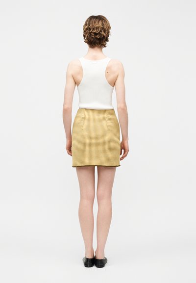 Άτομο με κοντά σγουρά μαλλιά, που φοράει λευκό αμάνικο τοπ, κίτρινη καρό φούστα και μαύρα παπούτσια, στέκεται πλάι με την πλάτη γυρισμένη προς λευκό φόντο.