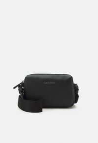 Bolso negro rectangular tipo bandolera hecho de material sintético suave con cierre de cremallera y correa ajustable. Presenta un logo de Calvin Klein.