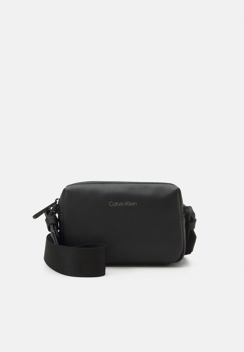 Bolso negro rectangular tipo bandolera hecho de material sintético suave con cierre de cremallera y correa ajustable. Presenta un logo de Calvin Klein.