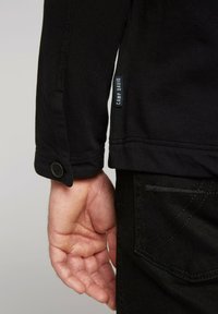 Giacca nera in tessuto strutturato con polsini abbottonati, dotata di un piccolo etichetta "CAMP DAVID" sulla manica. Jeans neri con cucitura sui taschini posteriori.