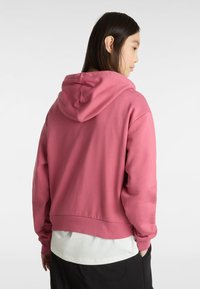 Rosa croppad huvtröja med huva, ribbat nederkant och långa ärmar. Slät textur, avslappnad design, bärs över en vit skjorta.