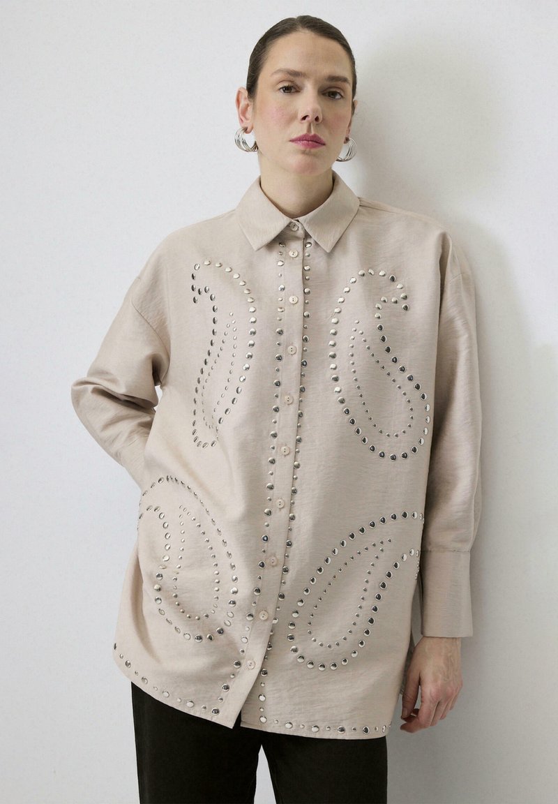 Beige skjorte med knapper og krave, prydet med metalnitter i paisley-mønstre på forsiden og brede ærmer. Glat, struktureret stof.