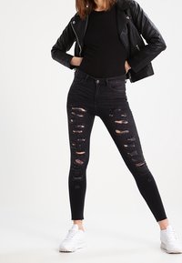Jeans skinny negros rasgados con múltiples parches desgastados y una cintura alta, combinados con una camiseta negra y una chaqueta de cuero, usados con zapatillas blancas.