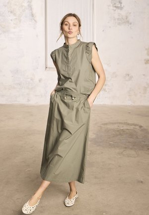 Femme en robe sans manches vert olive avec poches, debout sur un pied, faisant une moue de baiser, dans une pièce aux tons neutres avec des murs usés.