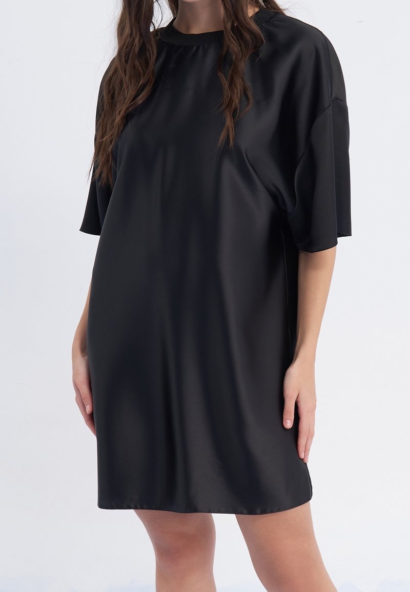 Femme portant une robe noire ample à manches courtes, debout avec les mains détendues le long du corps sur un fond uni.