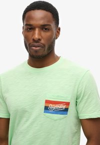 Camiseta de manga corta verde claro con una tela texturizada. Presenta un bolsillo en el pecho en colores contrastantes: rojo, naranja y azul con texto del logo.