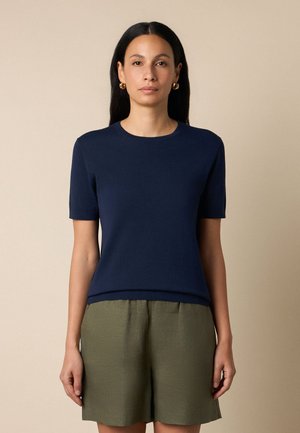 Frau trägt ein marineblaues Kurzarmoberteil und olivgrüne Shorts, steht vor einem einfarbigen beigen Hintergrund und schaut nach vorne.