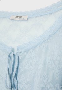 Jasnoniebieska koronka z motywami kwiatowymi, mała kokardka z przodu oraz metka z napisem "strteen" w pobliżu dekoltu.