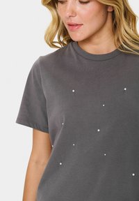Grå kortärmad t-shirt i mjuk bomull med små vita dekorativa pärlor jämnt fördelade på framsidan. Rund halsringning.