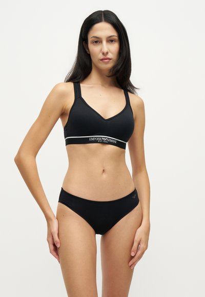 Femme portant un soutien-gorge de sport noir Emporio Armani et une culotte noire assortie, debout devant un fond blanc uni.