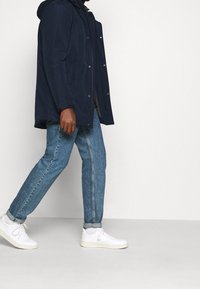 Person iført en marineblå jakke, lyseblå jeans med opsmøg, hvide sokker og hvide sneakers, der går imod en ensartet lys baggrund.