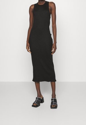 Czarna sukienka midi bez rękawów z gładkiej tkaniny, dopasowany krój o prostym sylwetce, w zestawieniu z masywnymi czarnymi sandałami z białym przeszyciem.
