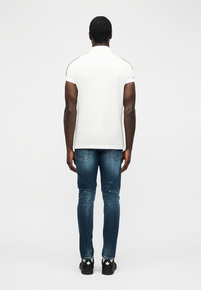 Homme debout, de dos, portant un polo blanc à manches courtes, un jean bleu avec des éclaboussures de peinture, et des baskets noires avec des logos blancs.