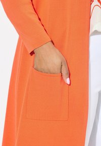 Orange kofta med en slät textur, som har en enda framficka och trekvartsärmar.