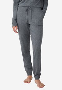 Persoon in grijze gebreide jogger broek met aantrekkoord in de taille en een cuff aan de enkels, staan met één hand in de zak, op blote voeten.