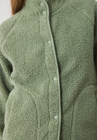 Fleece bunda v šalvějově zelené barvě, s měkkou a načechranou texturou, s patentkami a kontrastním zeleným lemem v oblasti límce a légu knoflíků.