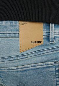Jeans de mezclilla azul claro con una cinturilla negra acanalada. Presenta una etiqueta de cuero marrón claro con la inscripción "CHASIN'" cosida en el bolsillo trasero.