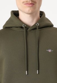 Olivengrønn hettegenser laget av myk stoff, med kengurulomme, justerbare snorer med sølvfarget metall og brodert logo.