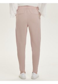 Ljust rosa tapered byxor med resårmidja, bärs med vita sneakers och en vit långärmad topp med spetsdetaljer vid ärmarna, visad bakifrån.